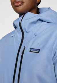 Αδιάβροχο μπουφάν ανοιχτού μπλε χρώματος με κουκούλα, με μαύρα φερμουάρ και το λογότυπο της Patagonia στο στήθος. Λείο υλικό, λειτουργικός σχεδιασμός.