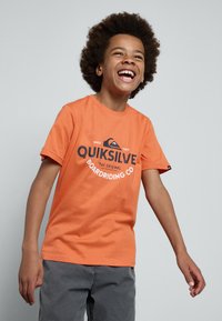Oranžové bavlněné tričko s černě potištěným logem "QUIKSILVER" a grafickým designem, s krátkým rukávem a klasickým kulatým výstřihem.