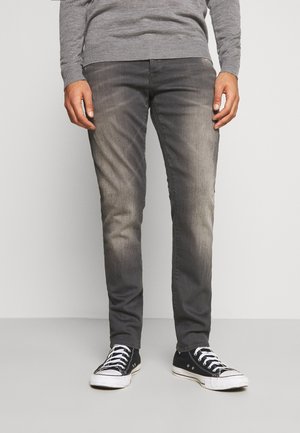 Homme portant un jean slim gris délavé, des baskets noires et blanches, et un pull gris à manches longues, debout sur un fond uni.