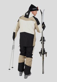 Lyžařský oblek v béžovo-černé barevné kombinaci, který sestává z bundy s kapucí a odpovídajících kalhot. Drží stříbrné lyže a lyžařské hole.