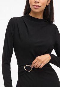 Femeie îmbrăcată într-o rochie neagră cu mânecă lungă, cu un guler înalt și o curea din material ce are o cataramă aurie neregulată.