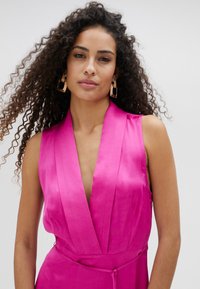 Vestido de satén fucsia con escote en V profundo y collar ancho; diseño sin mangas que presenta una cintura ceñida y textura suave. Pendientes de aro dorados.