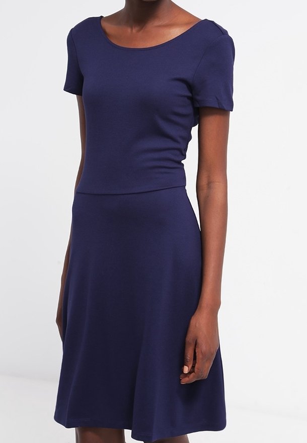 Femme portant une robe bleu marine ajustée à manches courtes avec un col rond et une jupe trapèze sur fond blanc uni.