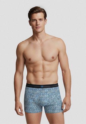 Uomo con capelli castani corti che indossa boxer blu con stampa leopardata e fascia nera in vita, in piedi su uno sfondo chiaro e uniforme.