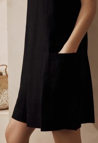 Une robe en lin noir avec une coupe décontractée, de longueur au-dessus du genou et des poches latérales. L'ourlet est légèrement courbé, avec une texture lisse.