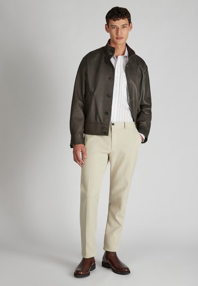 Brązowa skórzana kurtka z zapięciem na guziki, biały pasek na koszuli, beżowe spodnie oraz brązowe botki za kostkę. Gładkie faktury i dopasowany krój.