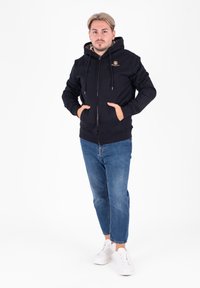 Felpa nera con zip e coulisse, dotata di tasche anteriori e foderata in plaid, abbinata a jeans blu e sneakers bianche.