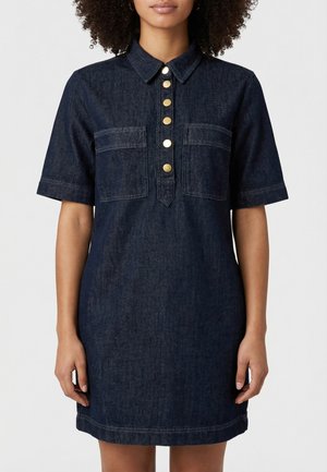 Vrouw draagt een donker denim jurk met korte mouwen, een kraag, twee borstzakken en een rij gouden knopen aan de voorkant.