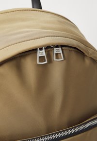Zaino beige con un tessuto testurizzato, dotato di una chiusura con zip superiore e doppie zip in argento. La parte inferiore presenta zip nere a contrasto.