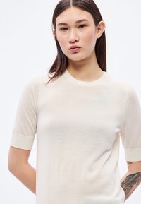 Jasnobeżowy sweter dziany z krótkimi rękawami, okrągłym dekoltem i subtelną fakturą. Tkanina wydaje się miękka i lekka.