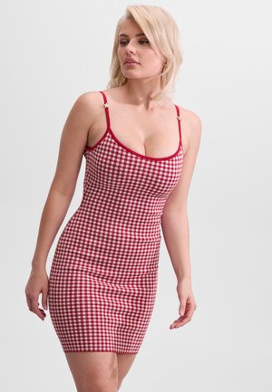 Blondynka ubrana w czerwoną i białą sukienkę w kratkę bez rękawów z cienkimi ramiączkami, lekko patrząca na bok na gładkim tle.