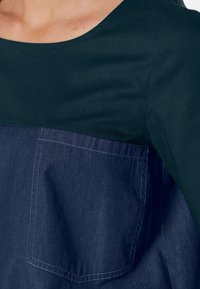 Maglietta a maniche corte con una sezione superiore verde scuro e una sezione inferiore blu navy, dotata di una tasca sul petto visibile e una texture simile al denim.