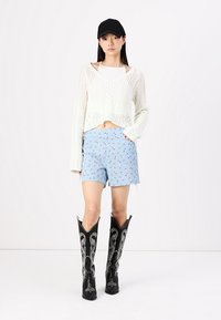 Vit stickad kort tröja, ljusblå mönstrade shorts och svarta cowboystövlar med silveraccenter. Modellen bär en svart keps.
