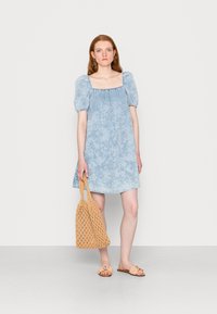 Vestido curto azul claro com decote quadrado, mangas bufantes e padrão floral. Bolsa de mão de palha bege e sandálias rasas completam o visual.