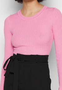 Růžové žebrované triko s dlouhým rukávem a kulatým výstřihem, nošené s černými kalhotami s vysokým pasem a zavazovacím páskem.