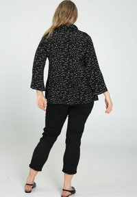 Camiseta negra con lunares blancos, de mangas largas y con cuello. Combinada con pantalones negros enrollados y sandalias negras. Fondo liso.