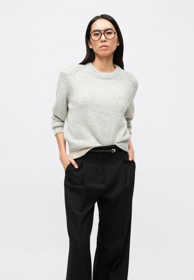 Sivý pletený sveter s rebrovanými detailmi, skombinovaný s čiernymi širokými nohavicami s prúžkami. Modelka má okuliare a ruky v vreckách.