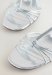 Ezüst színű magassarkú szandálok fényes, texturált felülettel. Keresztpántos kialakítás és karcsú sziluett jellemzi őket. Lapos talp, minimalista díszítések.