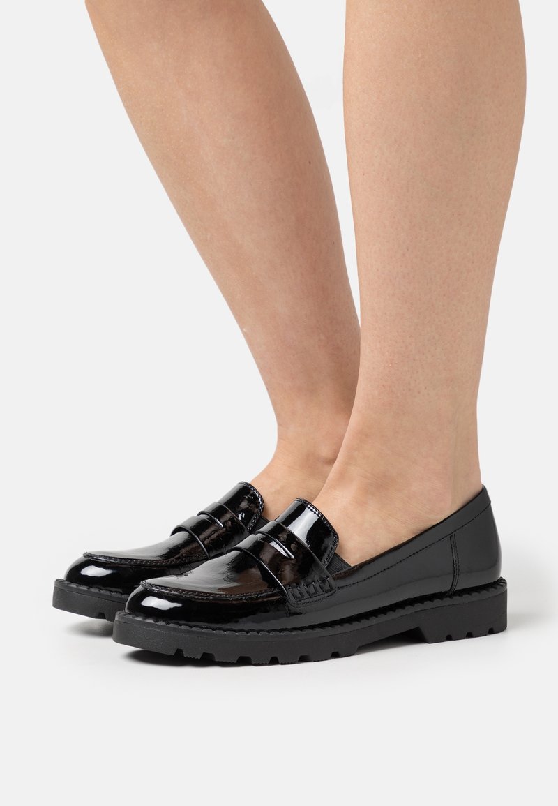 Jambes portant des mocassins en cuir verni noir brillant avec des semelles en caoutchouc épaisses sur un fond blanc.