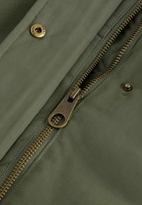 Olivgrüner Stoff mit glatter Textur, ausgestattet mit einem Reißverschluss aus goldfarbenem Metall und Druckknopfdetails an der vorderen Öffnung.