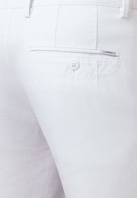Białe bawełniane spodnie o teksturowanym wykończeniu, z tylną kieszenią i zapięciem na guzik. Na kieszeni widoczna jest mała metka z logo marki.