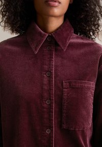 Bordeauxrode corduroy blouse met een puntkraag, knoopsluiting aan de voorkant en een borstzak. Voorzien van verticale ribbels en vier bijpassende knopen.