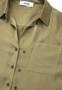 Camisa de botón verde oliva con una textura suave, cuello corto y un bolsillo en el pecho con botones a juego. Etiqueta de la marca: IPEKYOL.