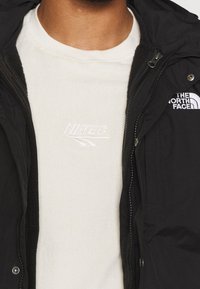 Personne portant une chemise blanche Hi-Tec partiellement recouverte par une veste noire North Face avec des logos visibles et des détails de fermeture éclair.