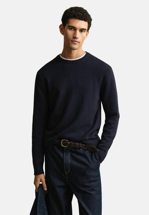 Young man with curly hair wearing a navy blue sweater, dark jeans, brown woven belt, holding a denim jacket in his left hand.