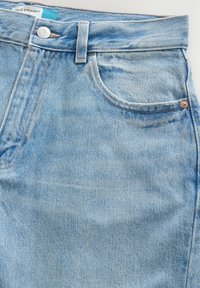 Gros plan sur un jean en denim bleu clair montrant la ceinture, le bouton métallique, la poche avant, la passants de ceinture et l'étiquette en tissu avec le texte "ANKLE STRAIGHT".