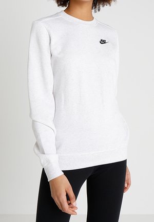 Vrouw draagt een effen witte Nike-trui met lange mouwen en een zwarte legging, staand tegen een neutrale achtergrond.