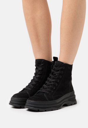 Bottes chelsea en suède noir avec une semelle en caoutchouc épaisse, présentant des lacets à l'avant et une boucle à l'arrière pour un enfilage facile.