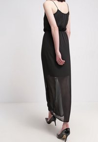 Černé maxi šaty s ramínky, průhledným dolním panelem a přiléhavým živůtkem. Nošeny s špičatými černými podpatky. Hladká textura, minimalistický design.