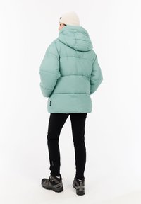 Persona de espaldas vistiendo una chaqueta acolchada verde menta con capucha, pantalones negros, zapatos de senderismo marrones y un gorro de punto blanco.