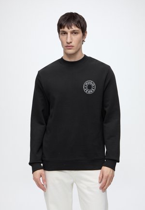 Giovane uomo che indossa una felpa nera Hugo Jeans con logo sul petto e pantaloni bianchi, in piedi davanti a uno sfondo bianco semplice.