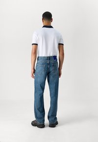 Muž stojící zády, oblečený v bílém polo tričku s černým lemem, modrých džínách a černých teniskách, na jednobarevném bílém pozadí.