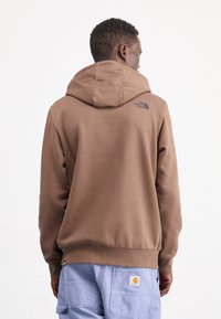 Bruine hoodie van zacht materiaal met een kangoeroezak. Bevat een logo op de achterkant en een capuchon met trekkoord. Gekozen in combinatie met lichtblauwe cargobroek.