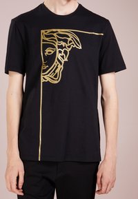 Camiseta de algodón negra con un diseño gráfico dorado de una cara y un acento de línea en el frente, con mangas cortas y un corte holgado.