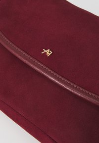 Pochette en daim bordeaux avec une bordure en cuir texturé, ornée d'un accent en métal doré de logo et d'une fermeture à glissière lisse.