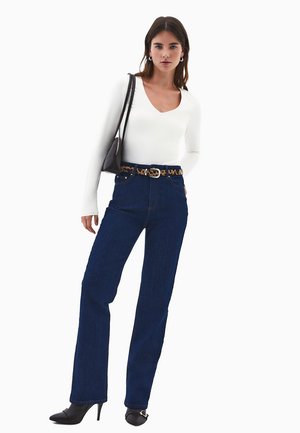 Donna che indossa una maglietta bianca a maniche lunghe con scollo a V, jeans blu scuro con cintura leopardata, scarpe con tacco nere e porta una borsa a spalla nera.
