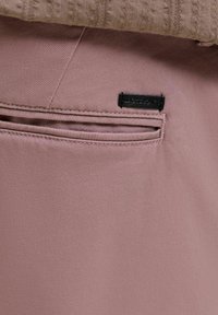 Lähivaade tagataskust tuhkurliivakarva pükstel, mille kohal tasku avamise kohal on must ristkülikukujuline brändi silt.