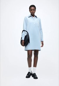 Šviesiai mėlyna suknelė su apykakle ir ilgomis rankovėmis, turinti užsegimą su sagomis ir kišenę. Derinama su juodais batais ir juoda rankine.