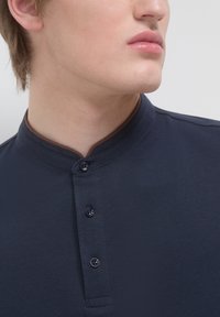 Polo shirt blu navy con tessuto testurizzato, dotato di colletto alla mandarino e tre bottoni scuri. Cuciture a contrasto marroni sul colletto.