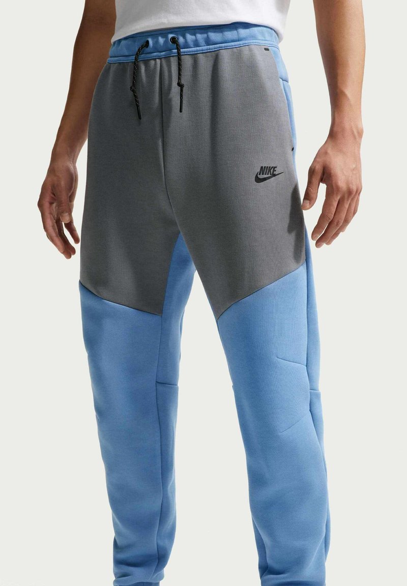 Pantaloni della tuta Nike con design a blocchi di colore, caratterizzati da tonalità grigie e blu, vita elastica con coulisse e tasche laterali.