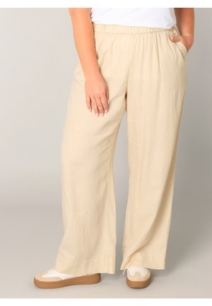 Persoon draagt losse beige broek met elastische tailleband en witte sneakers met rubberen zolen, staand tegen een effen achtergrond.