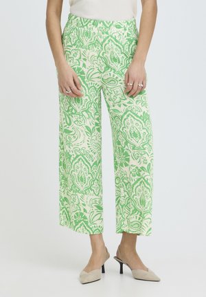 Kobieta ubrana w spodnie z wysokim stanem i szerokimi nogawkami w zielony kwiatowy wzór, biała bluzka, kilka pierścionków oraz beżowe sandały na obcasie, stojąca na białym tle.