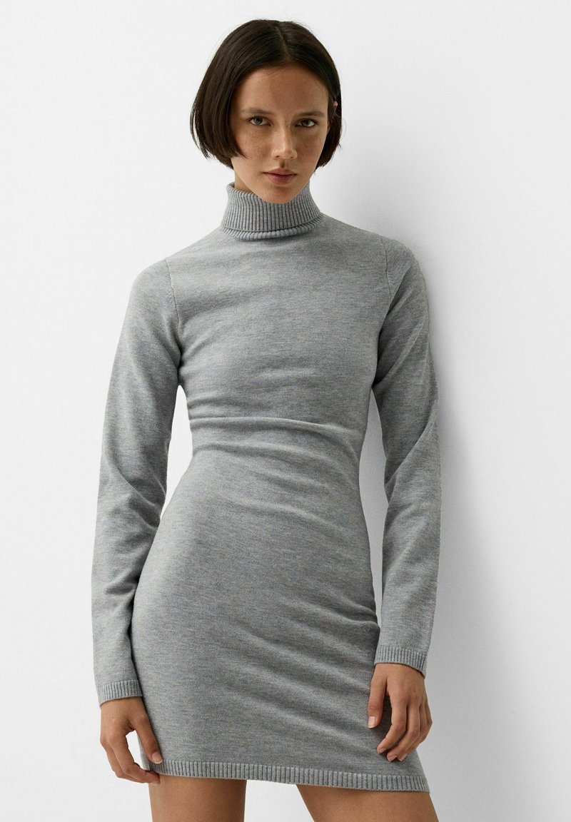 Frau mit kurzem dunklem Haar, die ein enges graues langärmliges Rollkragenstrickkleid trägt, steht vor einem schlichten weißen Hintergrund.