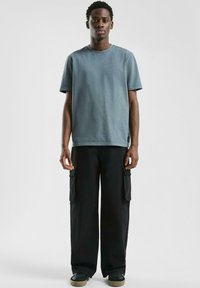 Jonge man staat gekleed in een blauw-grijze T-shirt met korte mouwen, zwarte cargobroek en zwarte schoenen met beige zolen voor een effen achtergrond.