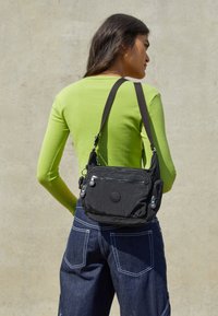 Černá nylonová křížová taška s dvěma zipovými kapsami, nastavitelným popruhem a detailem loga. Modelka má na sobě přiléhavé zelené tričko a modré denimové kalhoty.