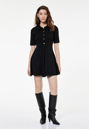 Kobieta ubrana w czarną sukienkę z krótkim rękawem zapinaną na guziki i czarne kozaki do kolan, stoi na tle jednolitego białego tła.